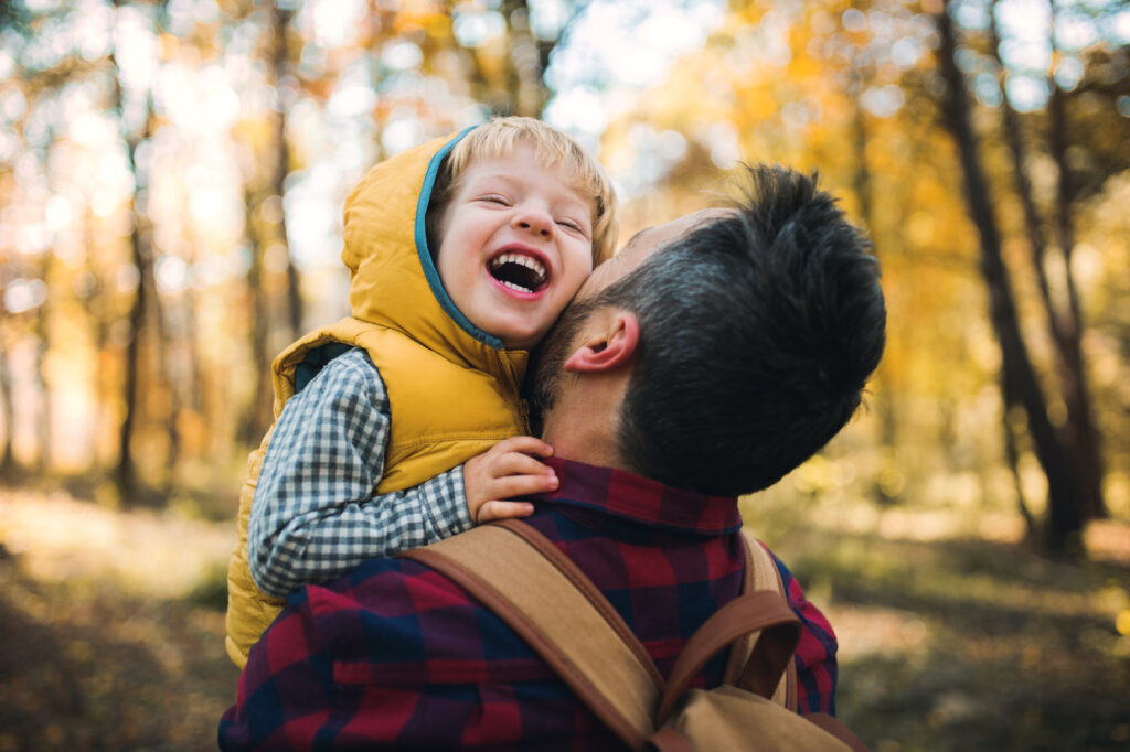 Ein Vater hält seinen jungen Sohn auf dem Arm. Der Sohn lacht. Sie sind im Wald spazieren.