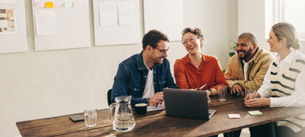 Ein gut gelauntes Team sitzt in einem Meetingraum an einem Tisch. Sie schauen auf einen Laptop, lachen und diskutieren miteinander.