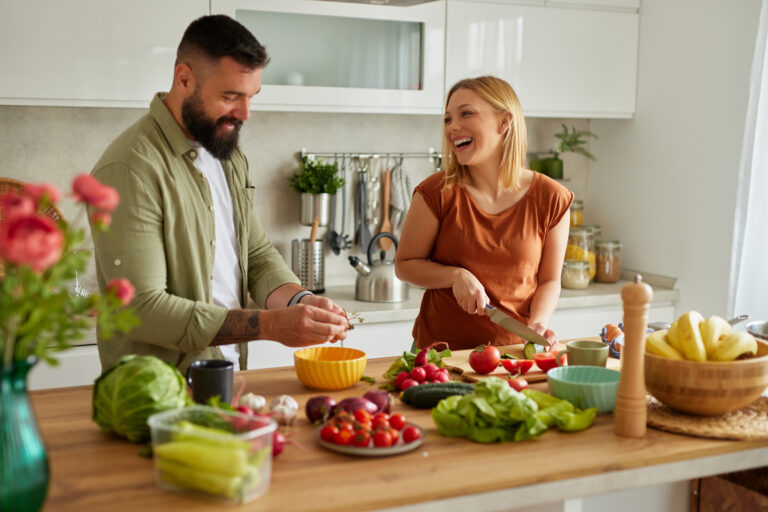 Ein junges Paar steht in der Küche und kocht gemeinsam. Auf der Küchenthrese steht verschiedenes Gemüse, das sie zubereiten. Sie sind gut gelaunt und lachen miteinander.