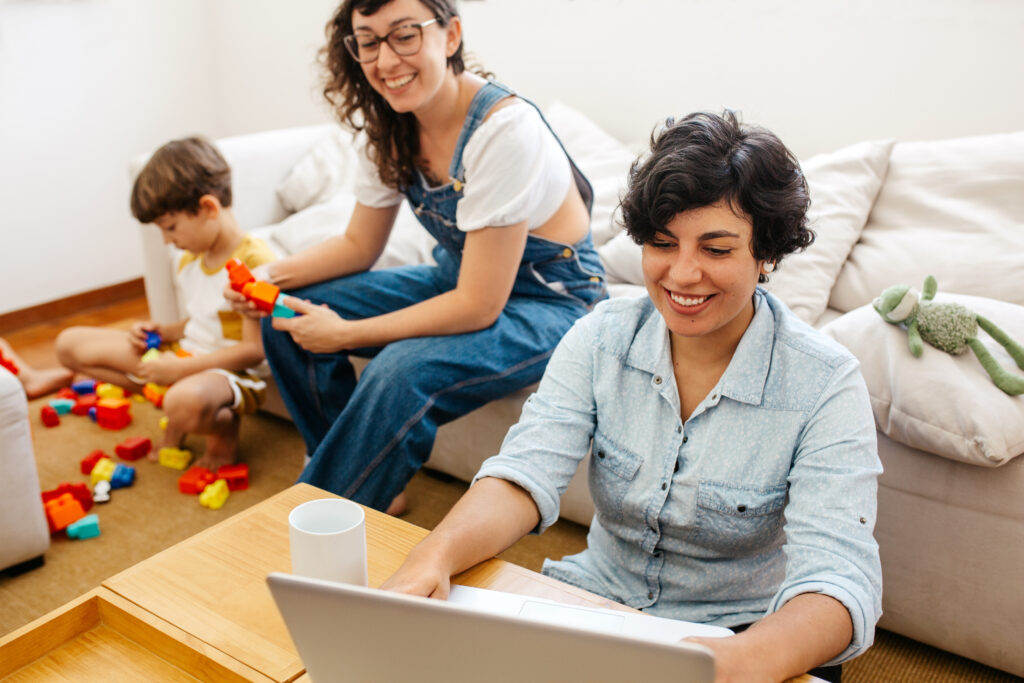 Eine Paar sitzt im Wohnzimmer. Eine Frau sitzt am Laptop. Die andere Frau spielt mit dem gemeinsamen Kind. Beide lachen.